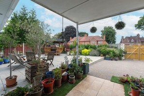 Ein überdachter Patio-Bereich mit Gartenmöbeln, Topfpflanzen und Blick auf Wohnhäuser.