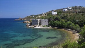 A coastal building with a stone facade, situated on a hillside with clear blue waters in the foreground.