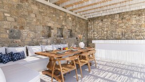 Un espace de restauration en plein air avec un mur en pierre en arrière-plan, des meubles en bois et un coin salon avec des coussins blancs.