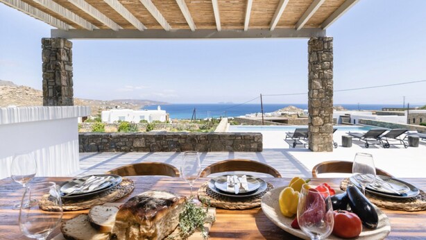 Une table dressée pour un repas, avec une vue sur la mer et des bâtiments blancs en arrière-plan.