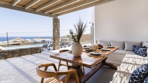 Une table à manger en bois, dressée avec des assiettes, un vase et des verres, donnant sur une piscine et offrant une vue sur la mer.