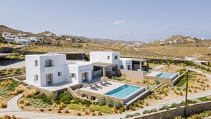 Une villa moderne aux murs blancs, dotée d’une piscine, entourée de jardins aménagés et située dans un paysage vallonné.