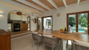 A modern kitchen with white cabinets, a wooden dining table, wicker chairs, and a view of a swimming pool. - Villa 'Marloria' with Mountain Views, Wi-Fi, and Air Conditioning (Crestatx)