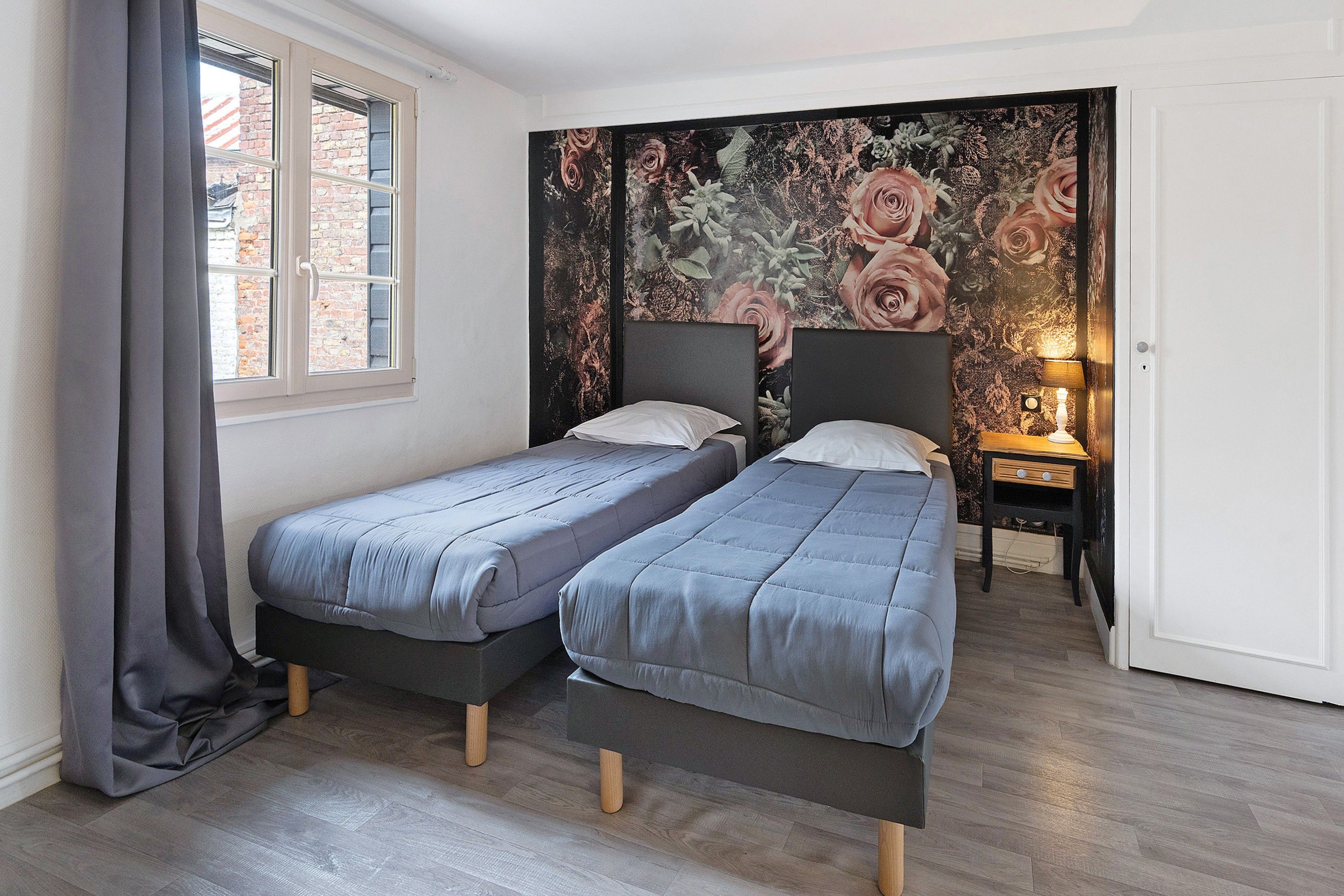 Ein Zimmer mit zwei Einzelbetten, einem fenster mit Vorhängen, einem Nachttisch mit Lampe und floralem Wandpapier.