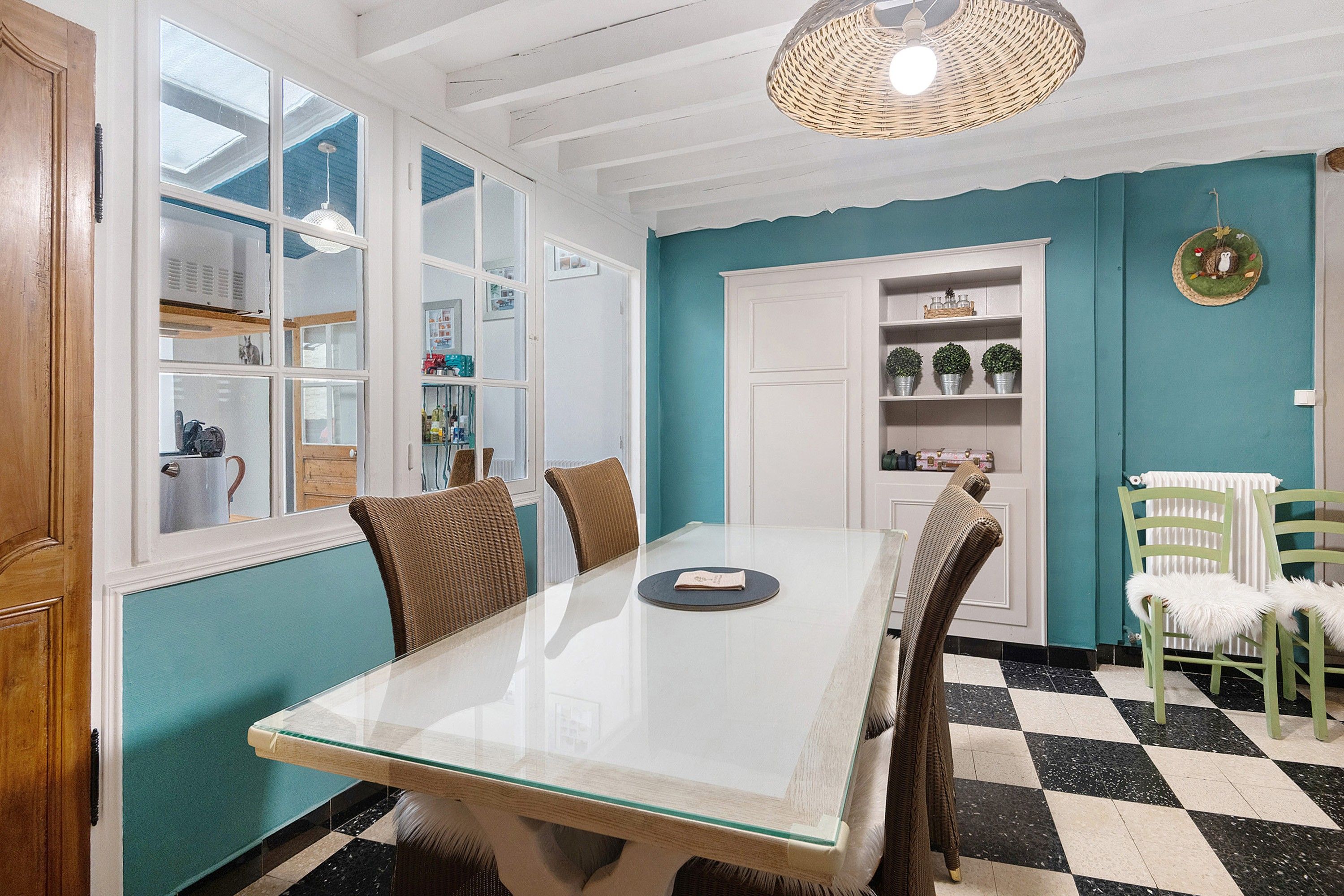A dining area with a glass table, wicker chairs, a white cabinet with glass doors, and a teal wall.
