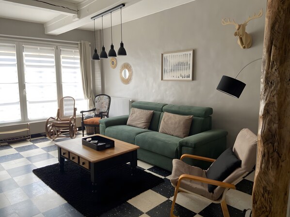 Un salon avec un canapé vert, une table basse en bois, un tapis noir, un sol à carreaux et une grande fenêtre avec des stores.