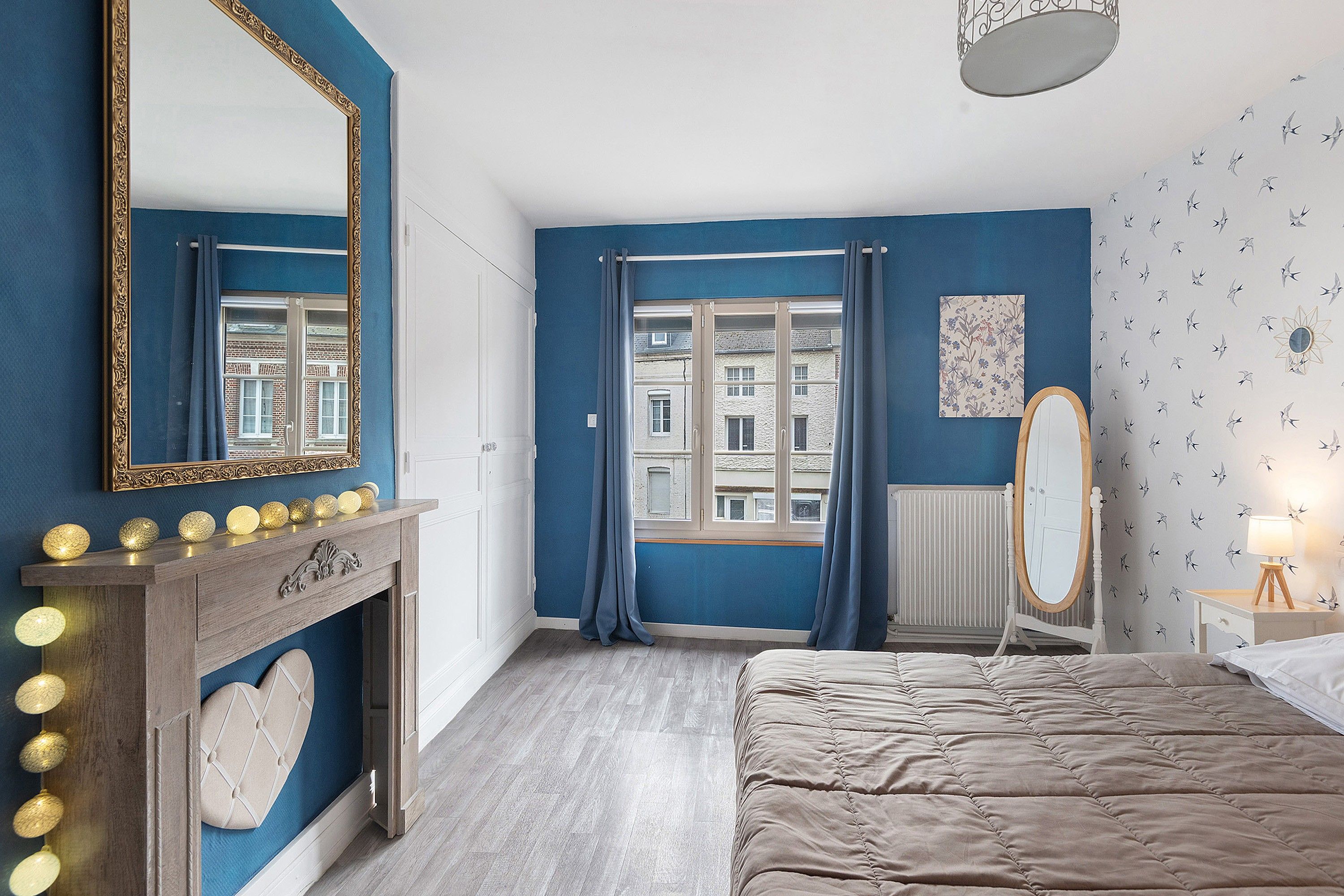 Une chambre à coucher avec un grand lit, une commode avec des lampes, un miroir, une fenêtre avec des rideaux et un radiateur.