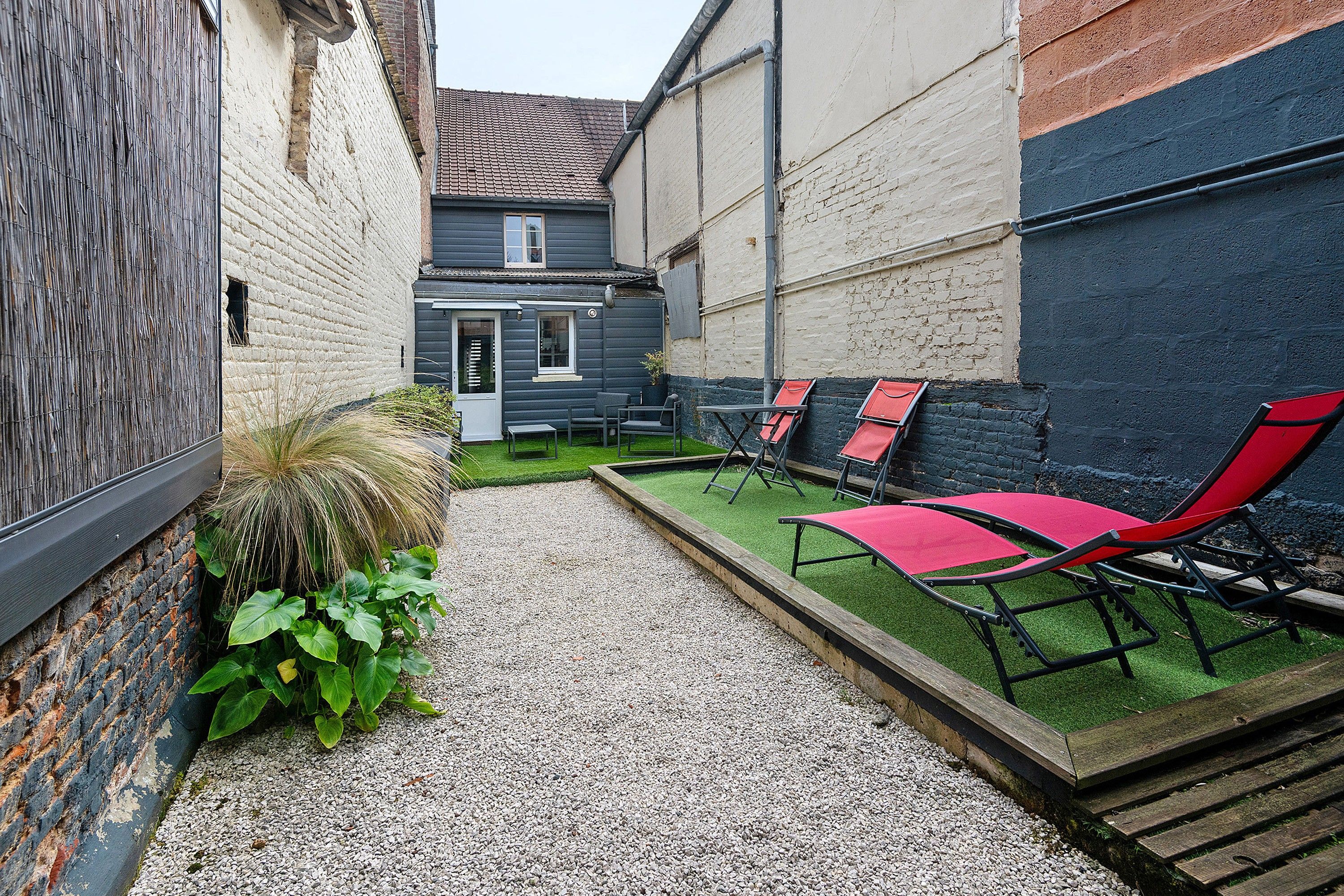 A narrow courtyard with a gravel path, a small garden area with plants, and outdoor seating with red chairs.
