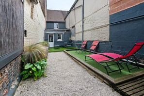 Ein schmaler Innenhof mit einem Kiesweg, einem kleinen Gartenbereich mit Pflanzen und Sitzgelegenheiten mit roten Stühlen.