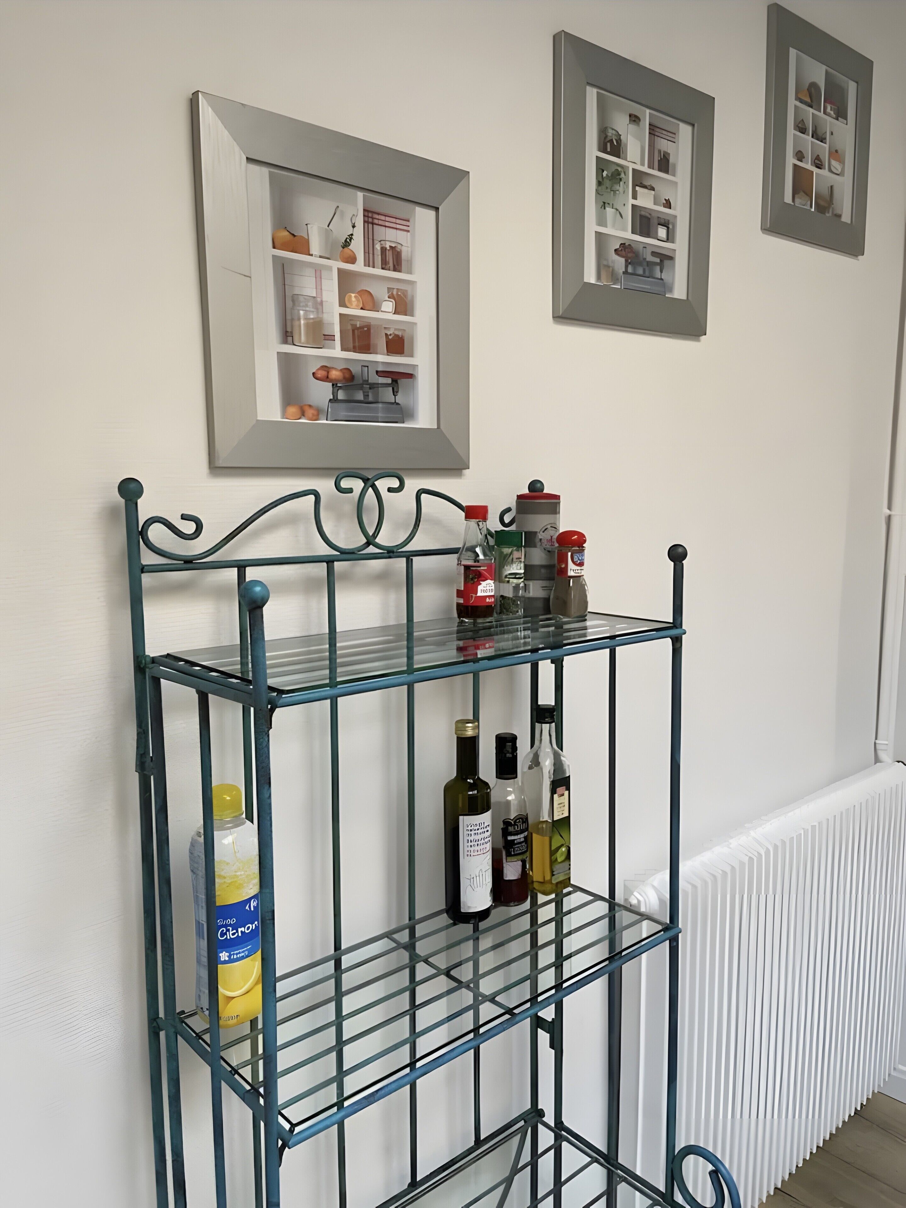 Une étagère métallique contenant des bouteilles et une boîte, appuyée contre un mur, avec des tableaux encadrés au-dessus.