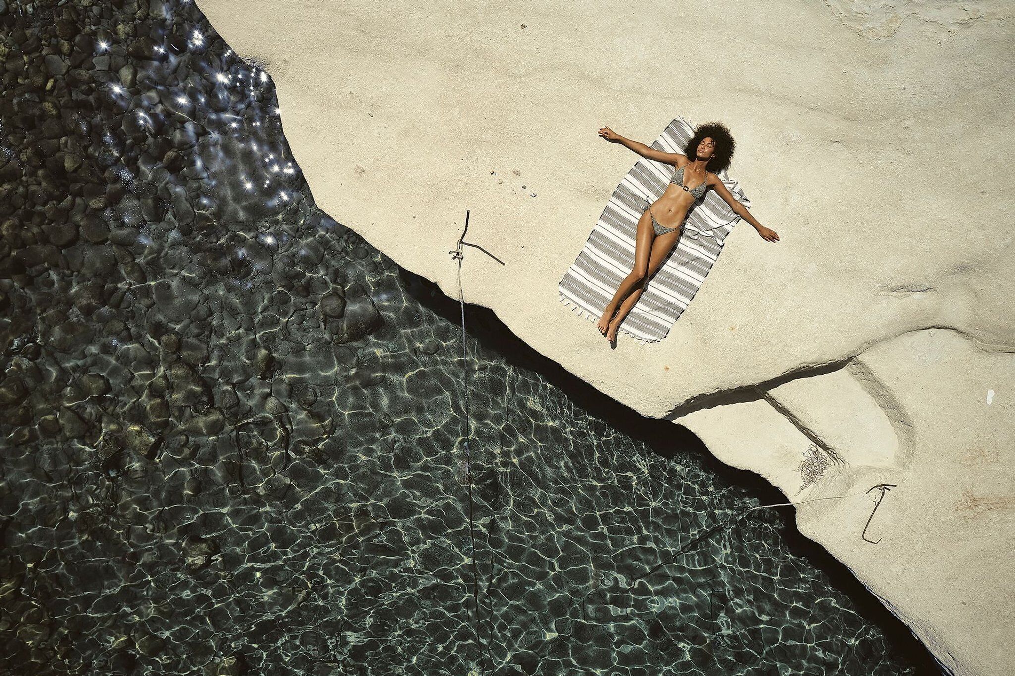 A person lying on a striped towel on a sandy beach with clear water.