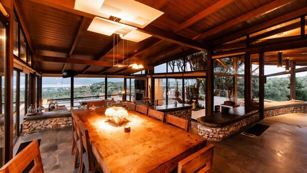 Un comedor amplio con una gran mesa de madera, detalles de piedra y ventanas grandes que ofrecen vistas del paisaje circundante.