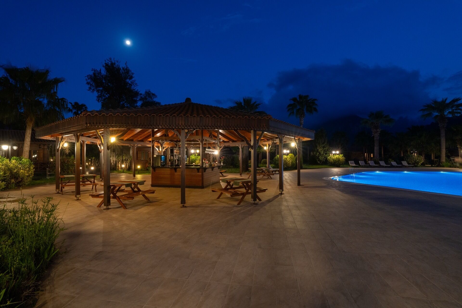 An outdoor pavilion with tables and benches, a pool, and palm trees at dusk.