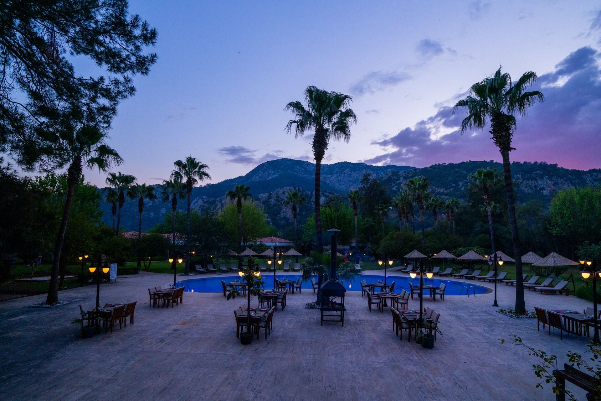 Área de piscina de un resort con sillas de descanso y sombrillas, rodeada de palmeras y montañas al fondo.