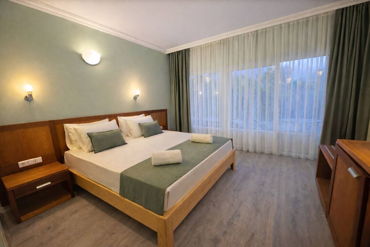 Ein Hotelzimmer mit einem hölzernen Bett, zwei Nachttischen, einem Schreibtisch und einem Fenster mit transparenten Vorhängen.