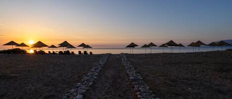 A beach with rows of umbrellas and lounge chairs, a pebble path leading to the water, and a sunset in the background.