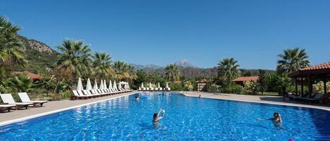 A swimming pool with lounge chairs and umbrellas, surrounded by palm trees and mountains in the background.