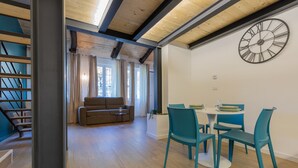 Ein modernes Wohnzimmer mit Sofa, Esstisch und Stühlen. Eine Treppe befindet sich links und eine große Wanduhr ist an der Wand.