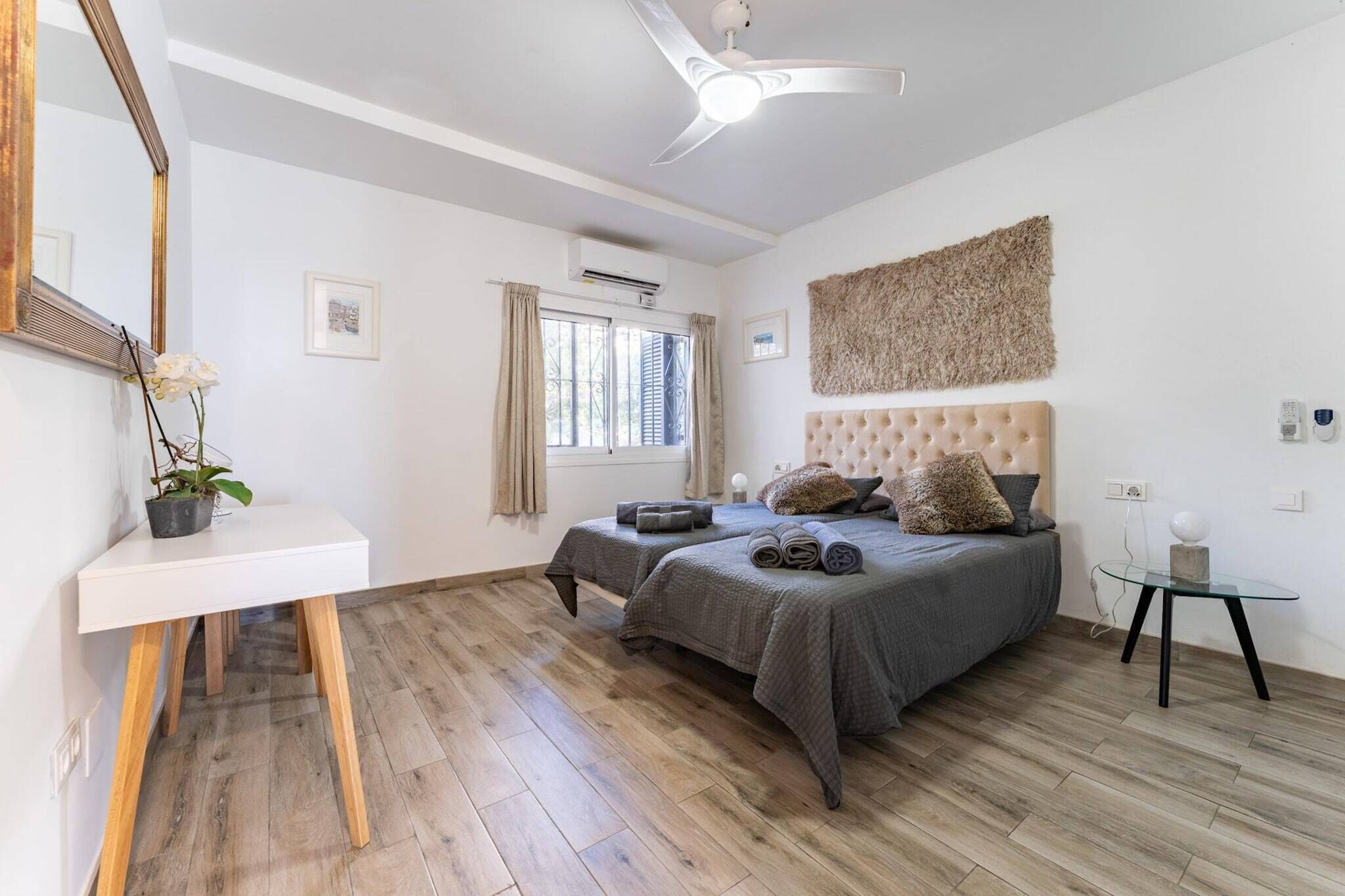 A bedroom with two beds, a white desk, a ceiling fan, and a large window with curtains.