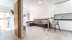 Ein moderner Wohnraum mit Ecksofa, zwei Barhocker, einem kleinen Tisch und einer Küchenzeile mit weißen Schränken und einer Marmor-Spritzschutzwand.