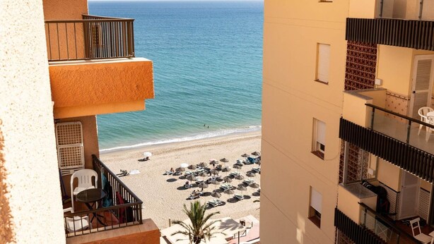 Ein Hochhaus mit Balkon mit Blick auf einen Strand mit Sonnenschirmen und Liegestühlen.