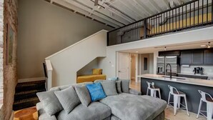 Un appartement moderne doté d’un grand canapé d’angle gris, d’une cuisine avec des meubles sombres et un réfrigérateur en acier inoxydable, ainsi que d’un escalier menant à un niveau supérieur.