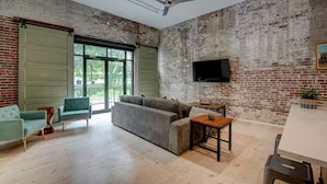 Ein geräumiges Zimmer mit großem Fenster, einer Couch, einem kleinen Tisch und einem Flachbildfernseher, der an der Backsteinwand angebracht ist.