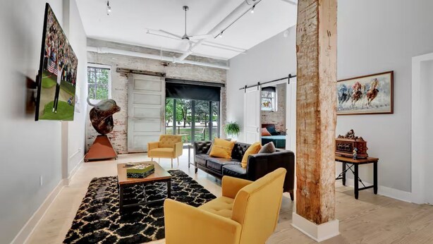 A spacious living room with a large sofa, yellow armchairs, and a coffee table. There is a painting on the wall and a sculpture on a pedestal. - The Goddard 1BR #102-Walking distance to downtown Hot Springs & 6 min to Oaklawn (Hot Springs)
