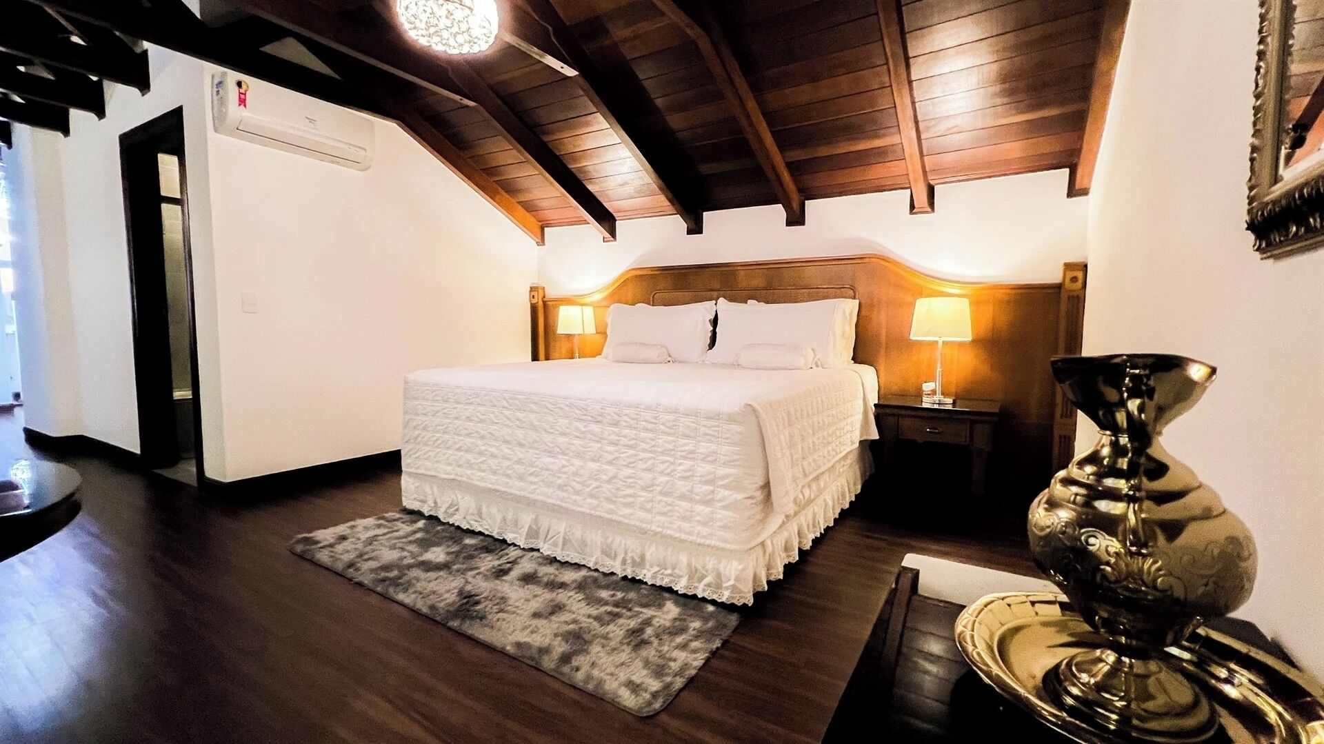 A bedroom with a large bed, wooden ceiling beams, a bedside table with a lamp, and a decorative vase.