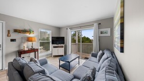 Una sala de estar moderna con un sofá gris, una mesita y un televisor. Hay un balcón con vistas al exterior.