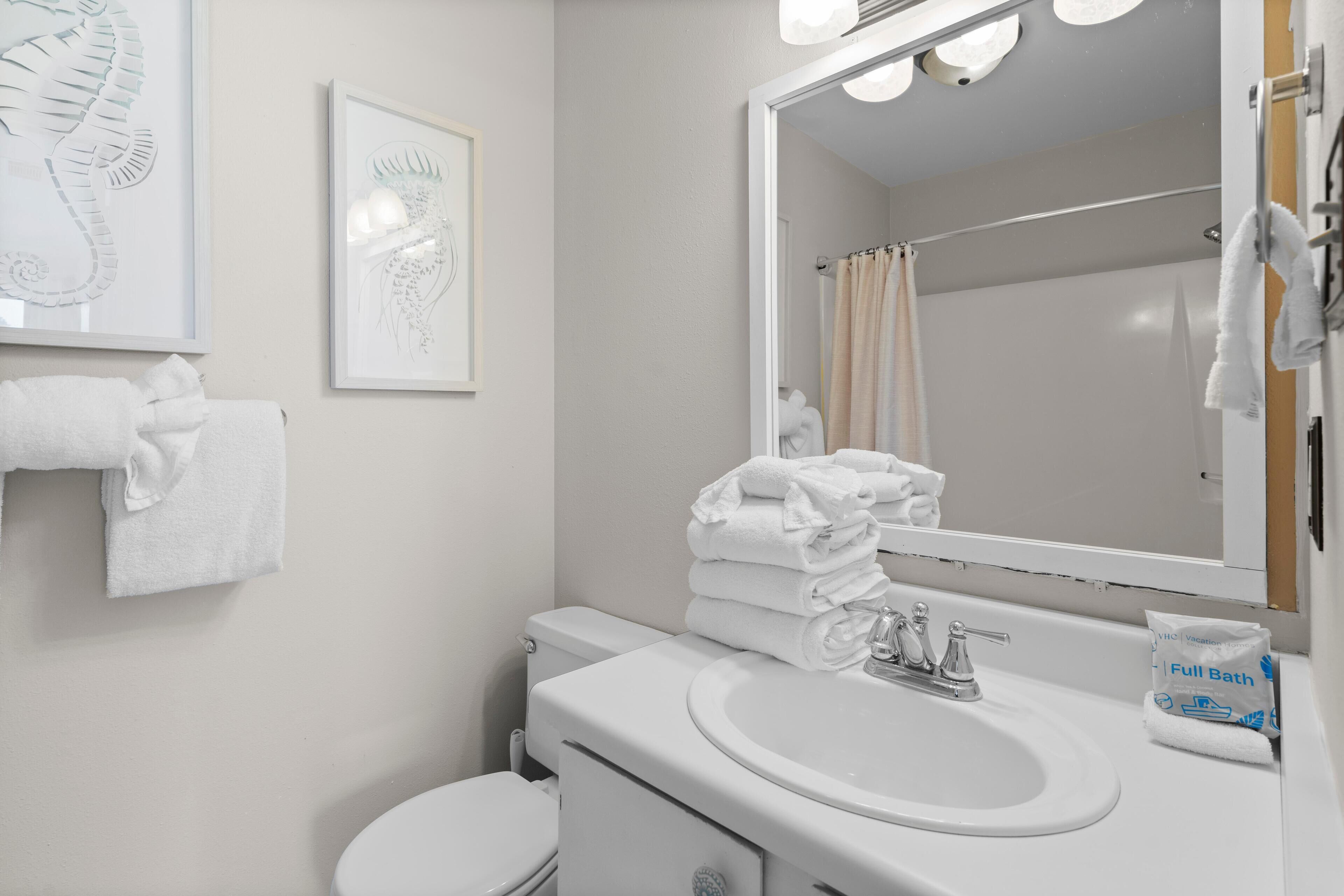 Un bagno moderno con lavandino, water e zona doccia, tutti bianchi. Asciugamani impilati sul lavandino e un quadro appeso alla parete.