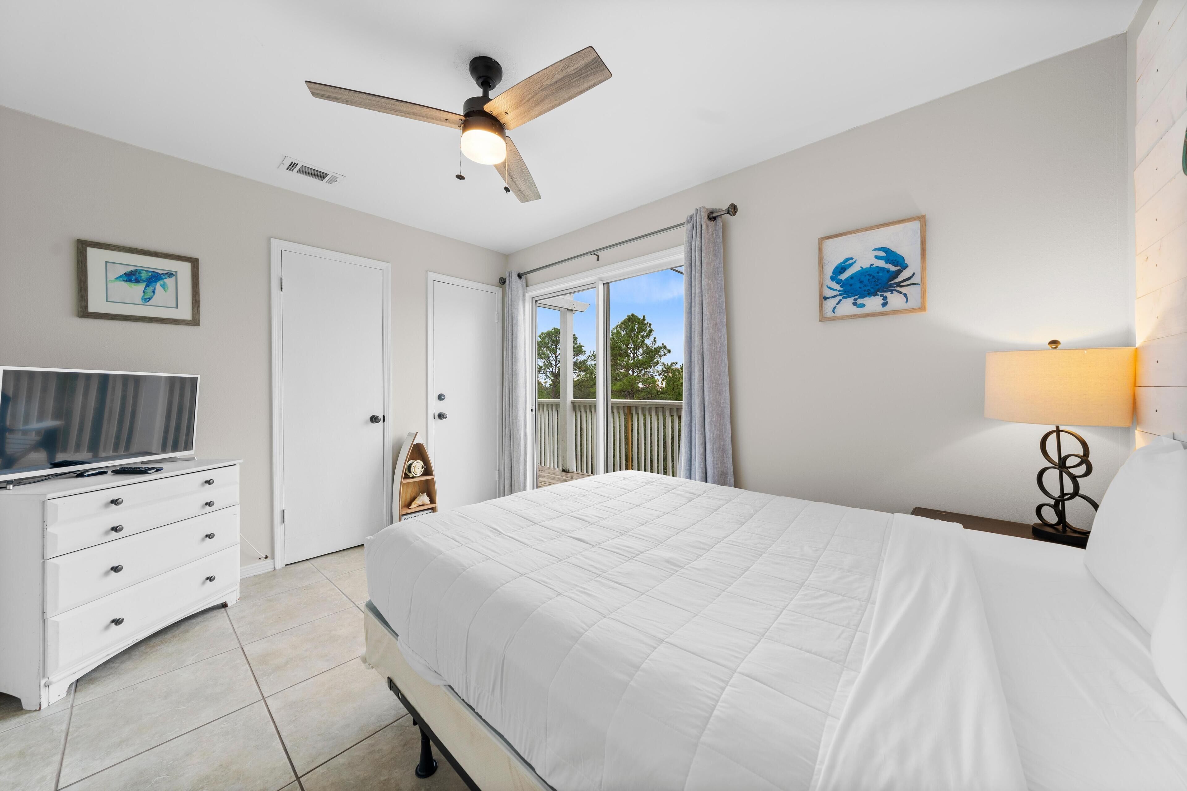 A bedroom with a bed, a TV, a ceiling fan, a window with curtains, and artwork on the walls.