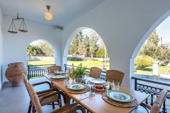 Un comedor con una mesa preparada para seis personas, rodeado de sillas de mimbre, con vistas a un jardín y árboles.
