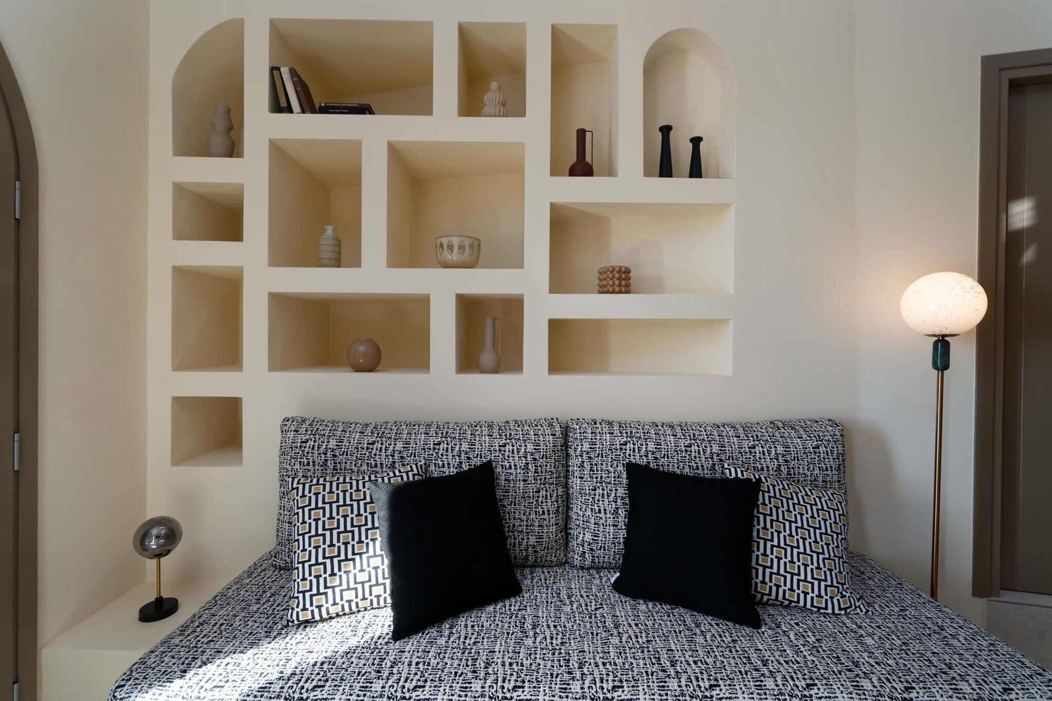 Um quarto com um edredom estampado, uma estante embutida com diversos objetos decorativos e um abajur.