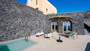 Zona de piscina con sillones reclinables y una mesa, paredes de piedra y vista al mar.