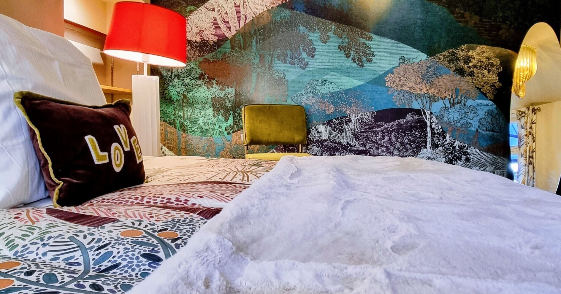 Een bed met een beddekleed met een patroon, een kussen met de tekst 'LOVE', een rode lamp en een groene stoel.
