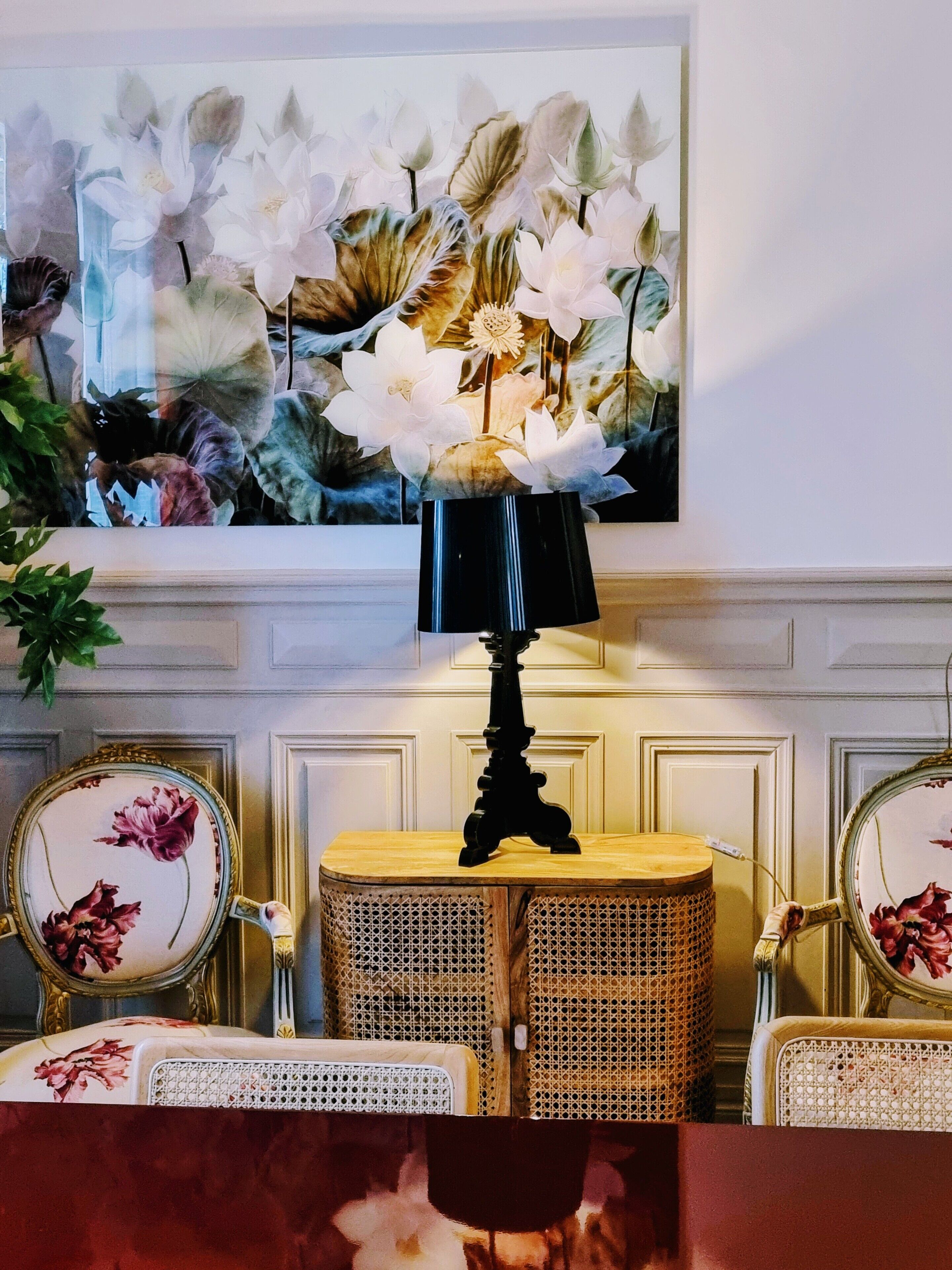 A room with a painting of flowers, a table lamp, wicker chairs, and a mirror with floral designs.