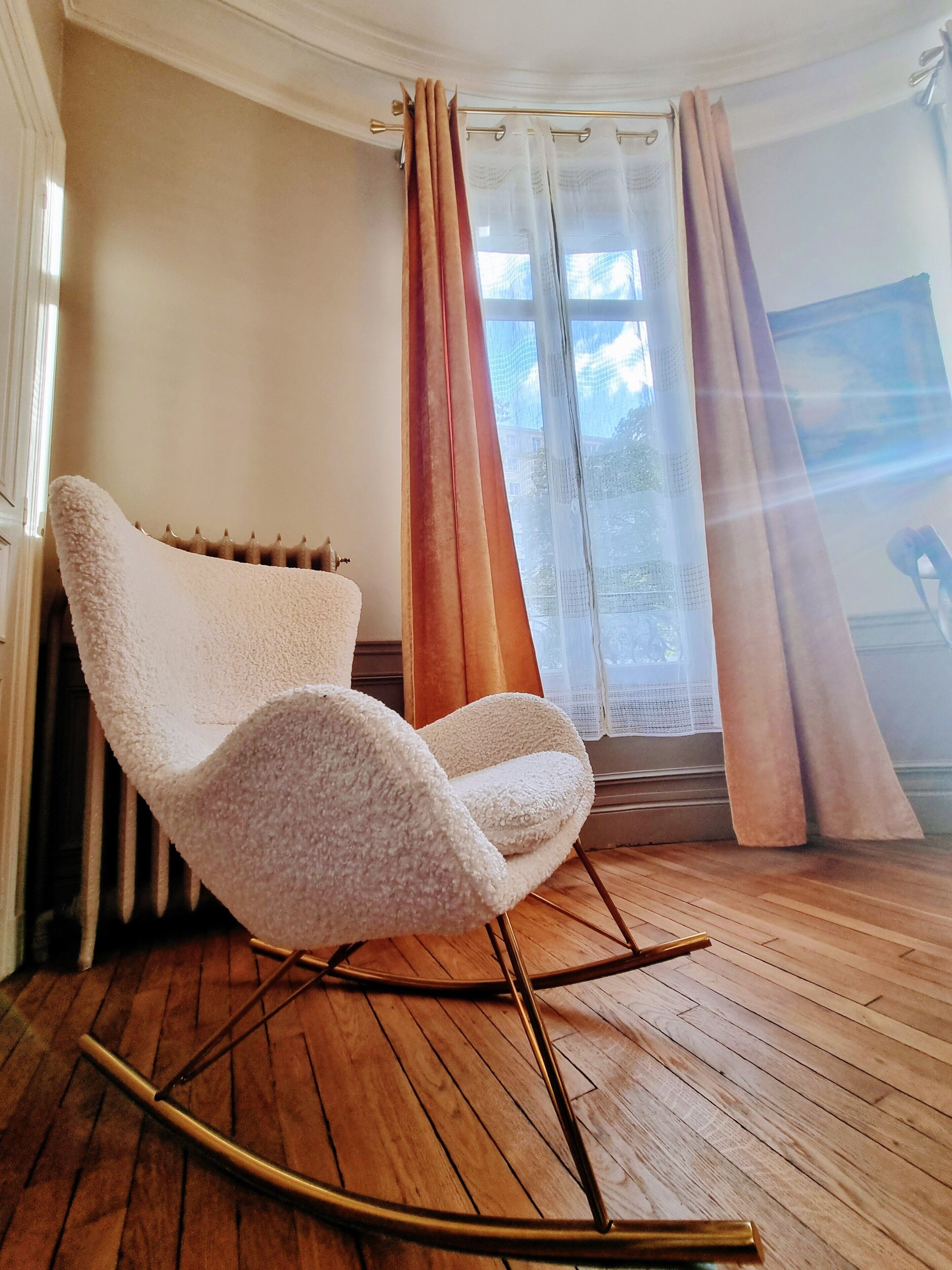 A room with a white textured chair, wooden flooring, a radiator, and a window with curtains.