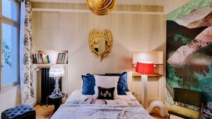 Un dormitorio con una cama, un espejo con forma de corazón, una araña, una estantería, una lámpara roja y un cuadro.