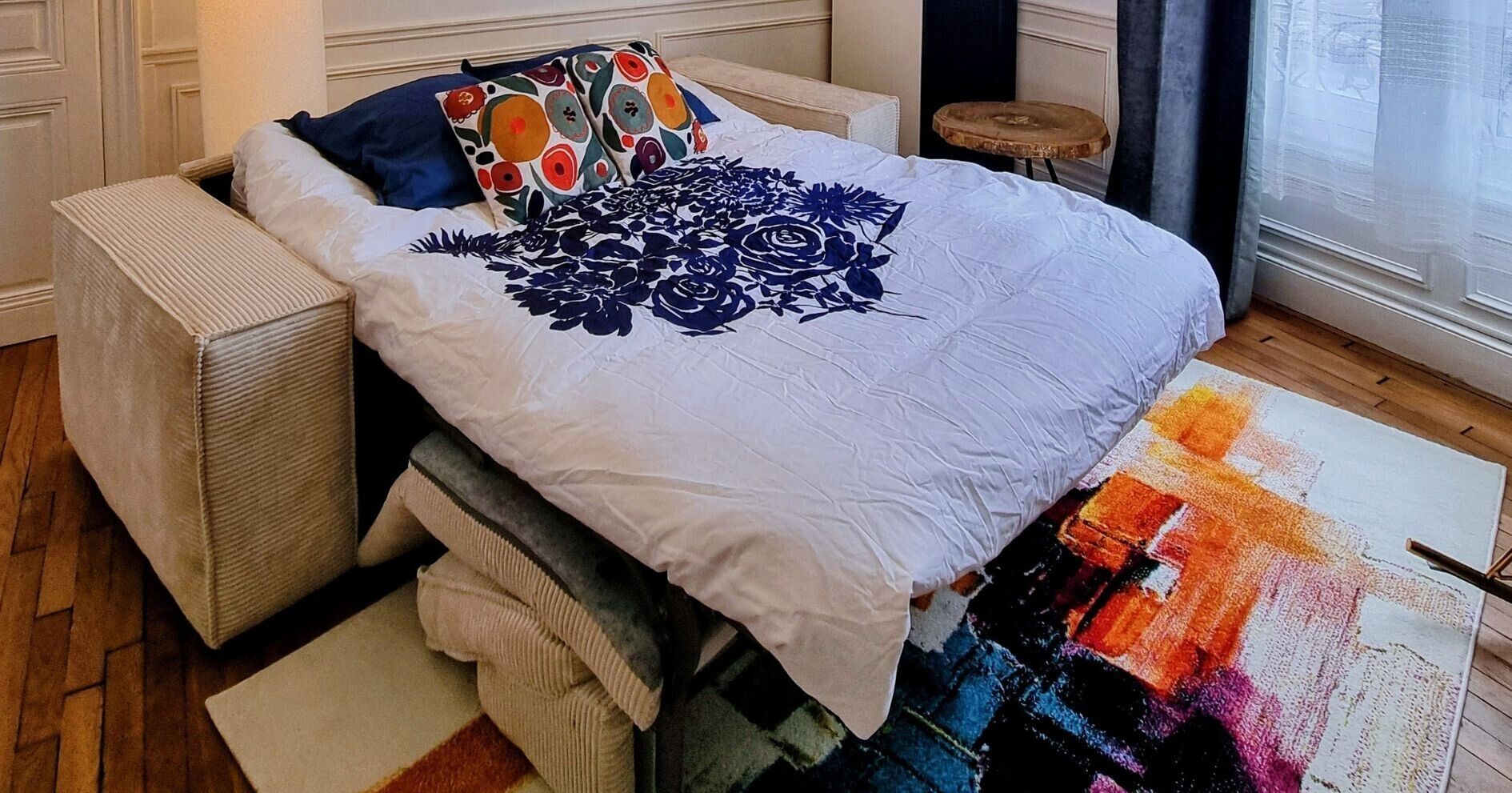 A bed with a floral bedspread, a colorful rug, and a small wooden stool.