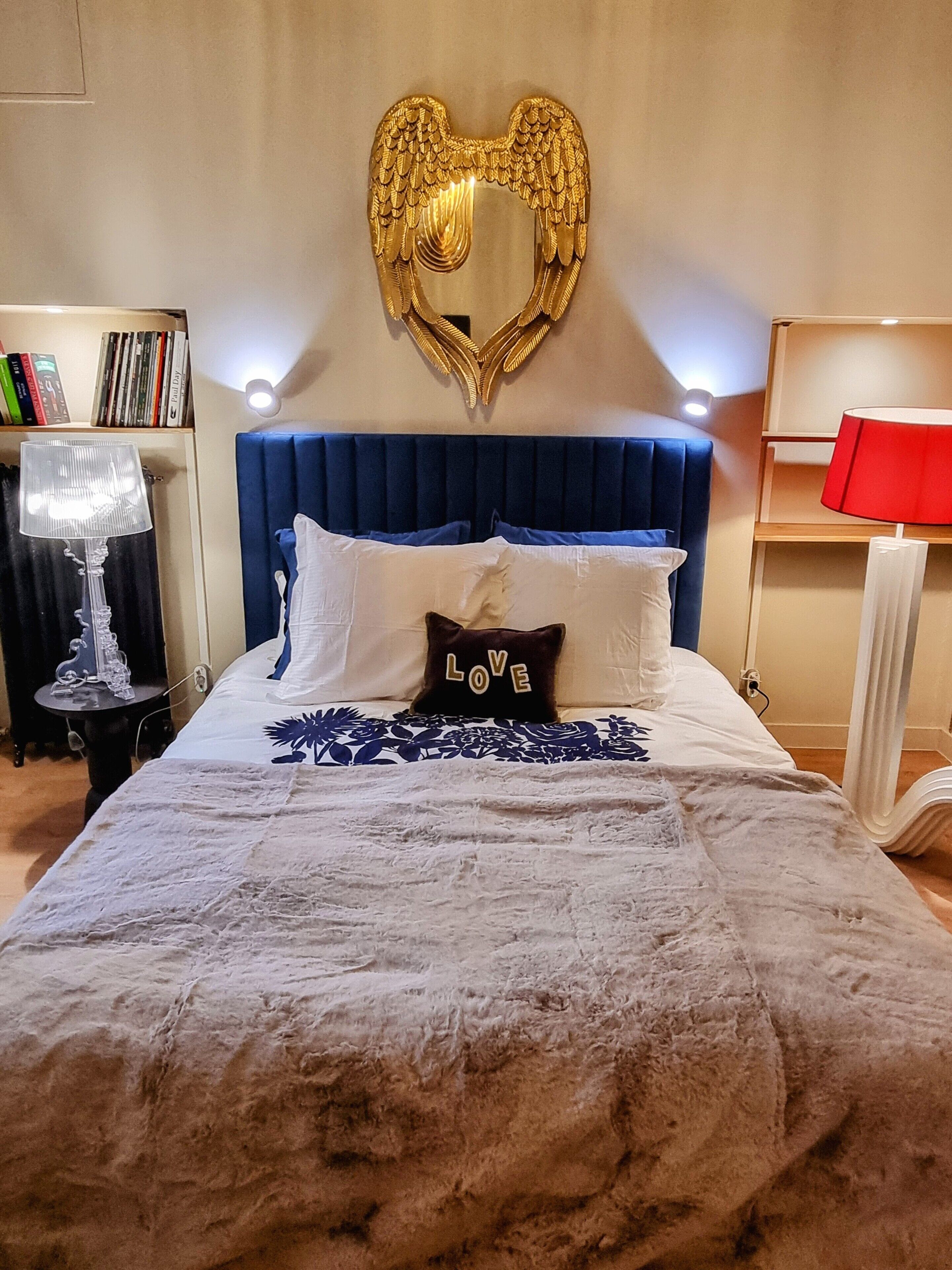 A bedroom with a heart-shaped wall decoration, a blue upholstered headboard, a white bedspread, and a pillow with 'LOVE' on it.