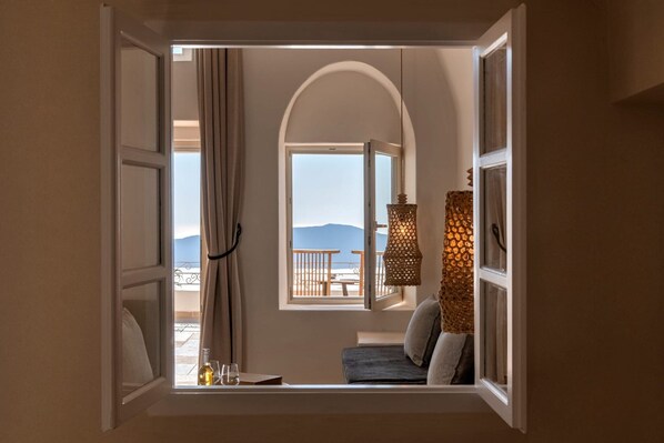 Una habitación con vistas al mar y a las montañas a través de una ventana abierta, un balcón con una mesa y sillas, y una lámpara colgante.