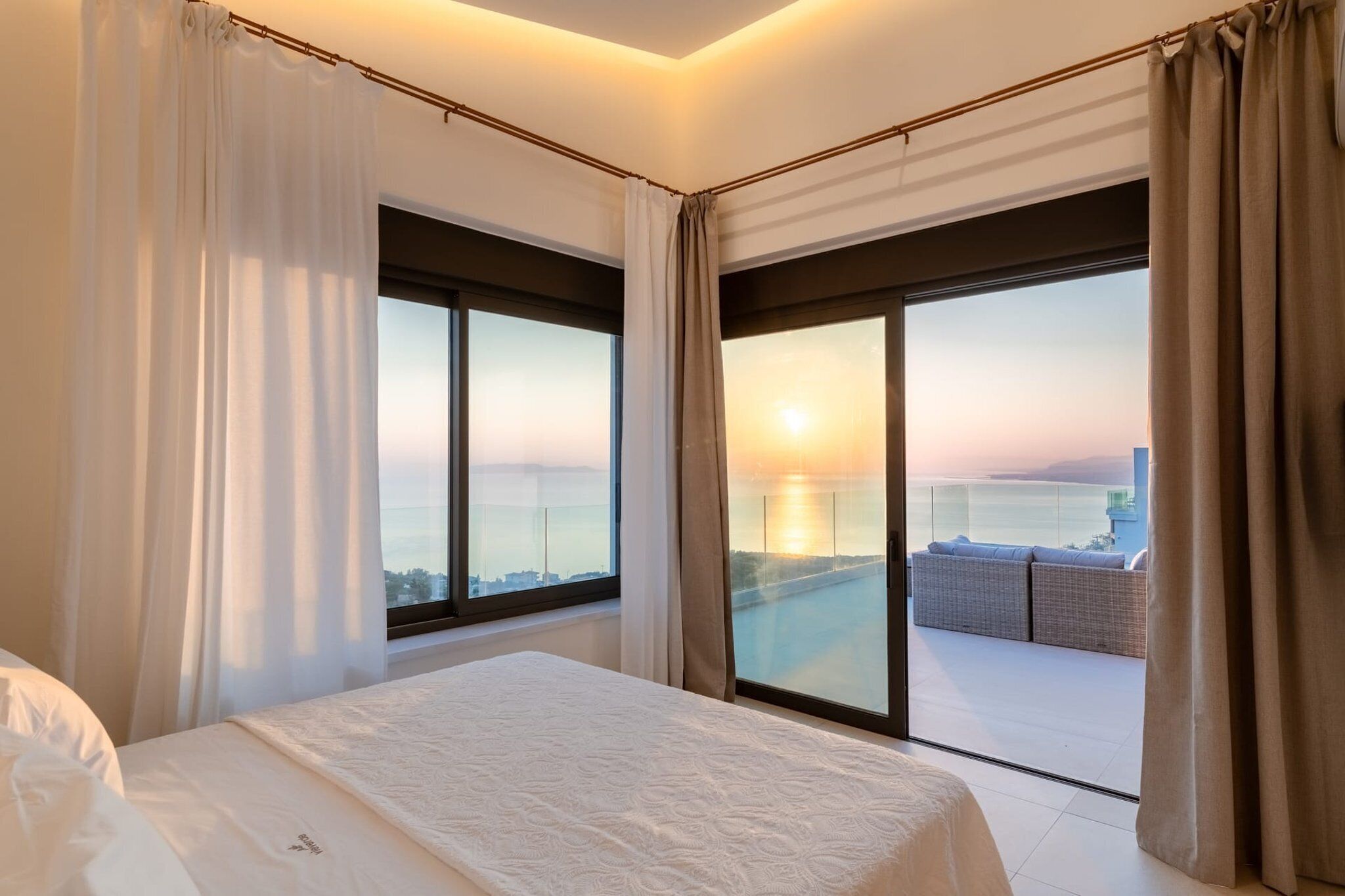 A bedroom with a large bed, two windows with curtains, and a view of a sunset over a body of water.