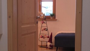 Een kamer met een houten deur, een bed met donkere dekbedovertrek, een klein tafeltje met een plant en een kerstboom met versieringen.