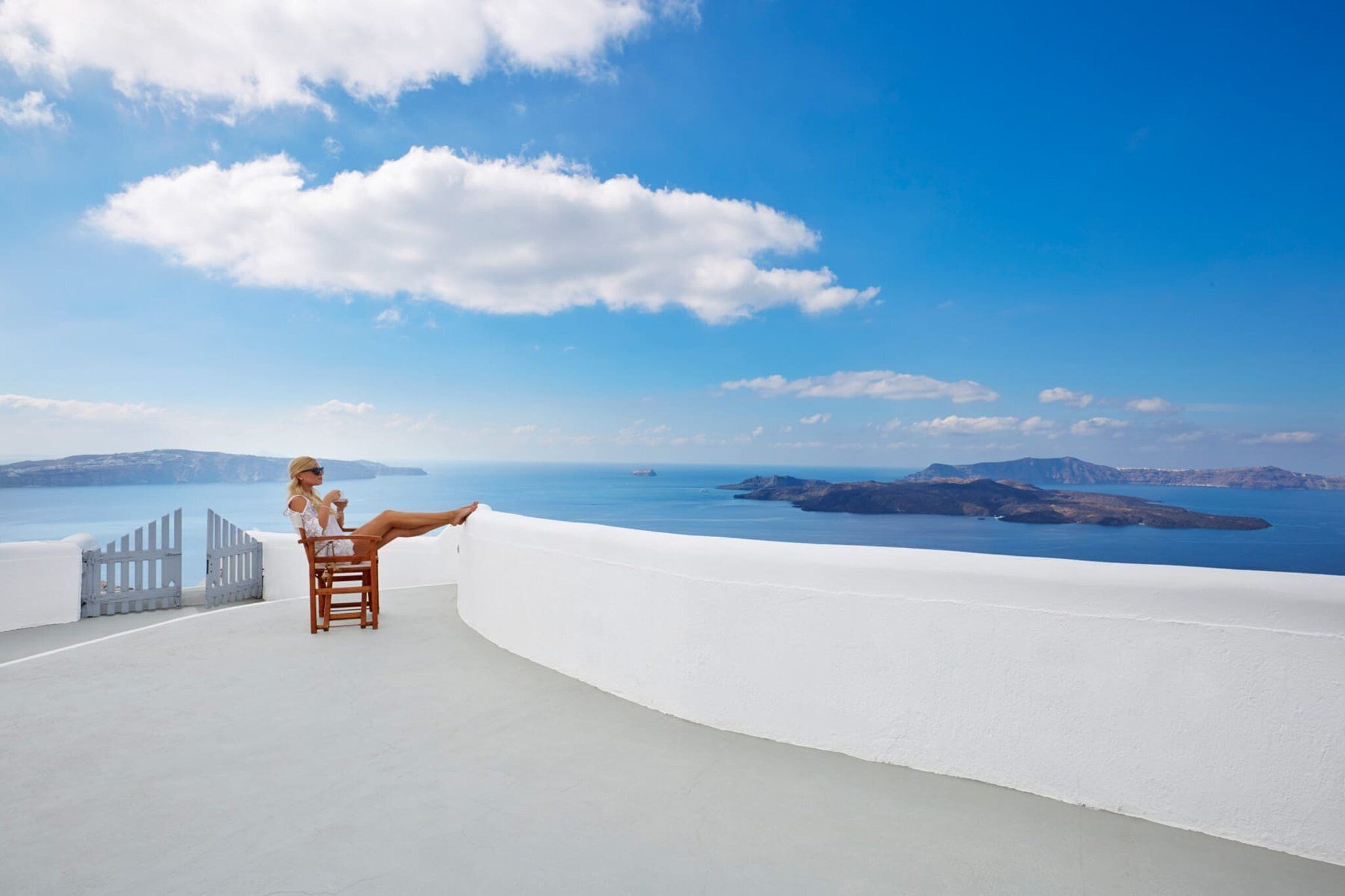A person seated on a white chair on a white balcony overlooking a coastal landscape with clear skies and distant islands.