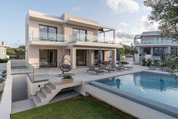 Een modern huis met twee verdiepingen, een zwembad, terrasmeubilair en een glazen balustrade.