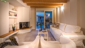 Un salon moderne avec un canapé blanc, une table basse et une cheminée. Le plafond est en bois et une porte vitrée offre une vue sur l’extérieur.