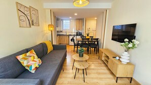 Ein modernes Wohnzimmer mit einem grauen Sofa, einem kleinen runden Tisch und einer Holzkonsole. Im Hintergrund ist eine Küche mit Essbereich zu sehen.