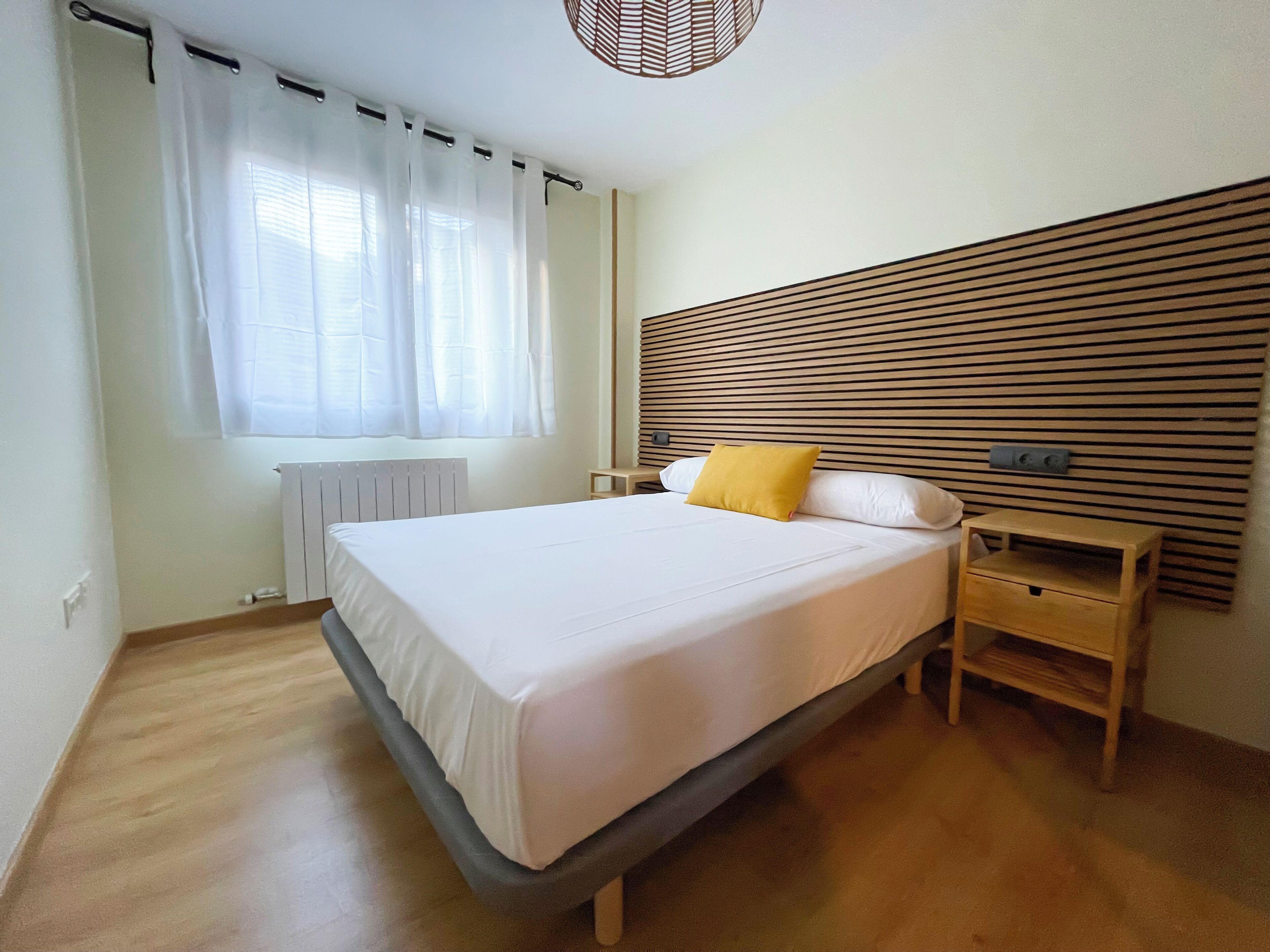 Een slaapkamer met een bed, nachtkastje, houten lattenborden hoofdeinde en een raam met gordijnen.