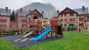 Ein Spielplatz mit einer blauen Rutsche und Klettergerüsten, umgeben von Steinhäusern mit Dachgauben, vor einer nebligen Bergkulisse.